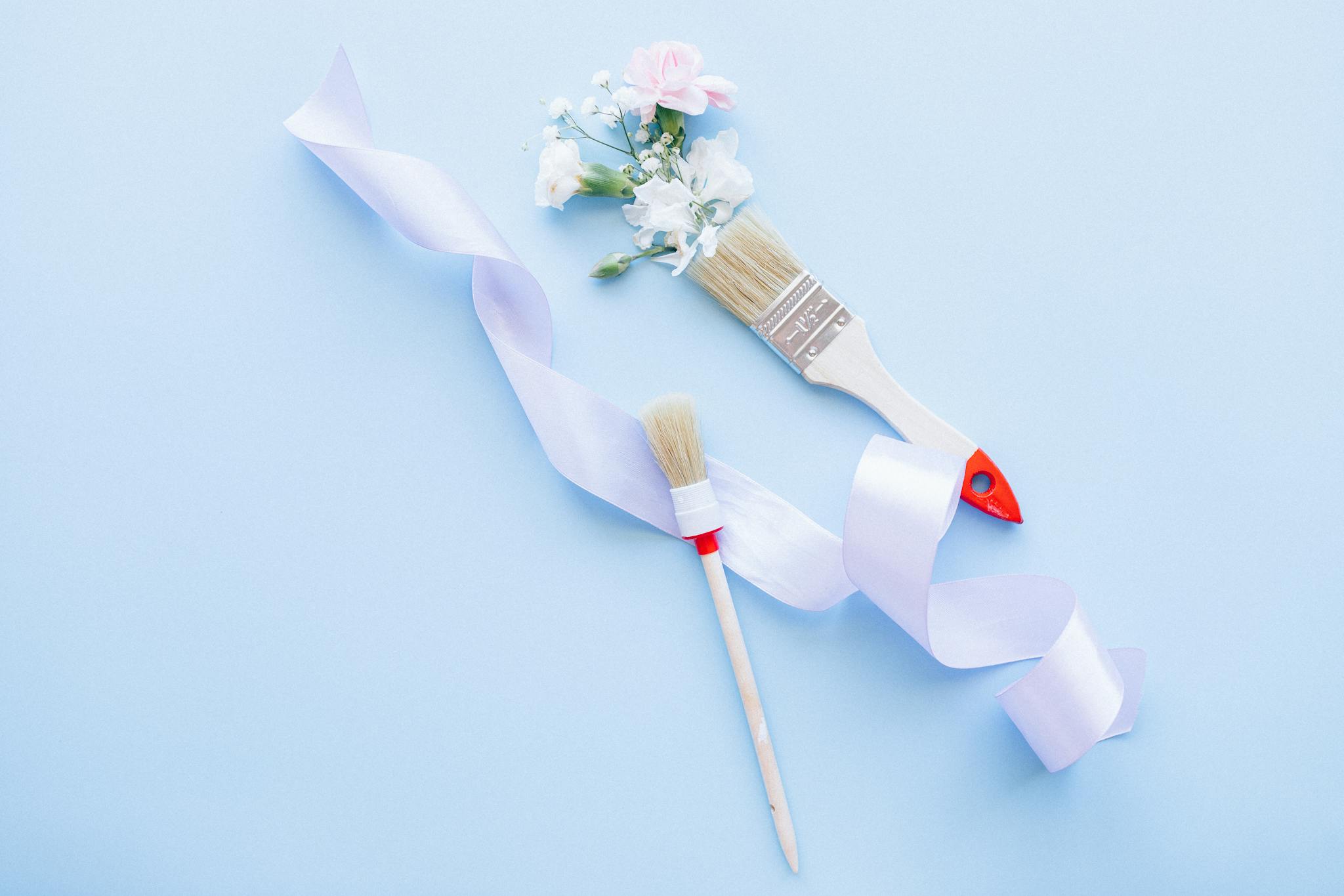 Two paint brushes with ribbon and flowers on a pastel blue background, ideal for DIY and decor themes.
